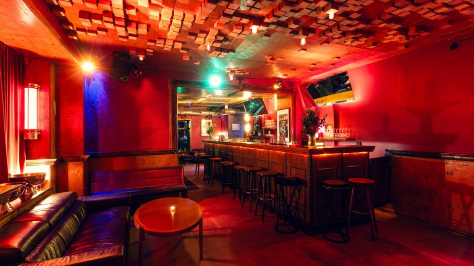 Innenraum einer Bar mit roter Wandgestaltung, langer hölzerner Theke mit Barhockern und gedämpfter, farbiger Beleuchtung. Markant sind die Decke mit akustischen Holzpaneelen, eine Sitzecke mit Sofa und runden Tischen sowie Wandmonitore und Spiegel hinter der Bar.