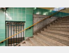 Treppenaufgang in einer U-Bahn-Station mit grünen, glänzenden Kacheln an den Wänden, einem gelben Handlauf und einem Geländer. Im Hintergrund ist eine Öffnung mit Blick auf eine Fliesenwand, auf der "Wittenbergplatz" steht.