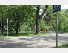 Straßenszene mit einem Fußgängerüberweg, flankiert von Bäumen und Grünflächen in einem parkähnlichen Umfeld. Im Hintergrund stehen eine Bank, ein orangefarbener Mülleimer und Verkehrsschilder, die auf die Straßenkreuzung hinweisen.