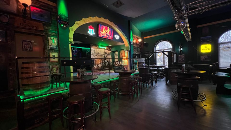 Innenraum einer irisch gestalteten Bar mit dunklen Wänden, hohen Tischen und Barhockern sowie Holzfässern als Stehtische. Im Hintergrund sind eine kleine Bühne mit beleuchtetem Torbogen, Leuchtreklamen und große Rundbogenfenster zu sehen.