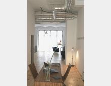 Helles, modernes Büro mit Holzboden, einem langen Holztisch und zwei Stühlen im Vordergrund. Im Hintergrund sind große Fenster, eine Sitzgelegenheit und sichtbare Lüftungsrohre an der Decke erkennbar.