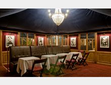 Ein eleganter, rot getönter Speiseraum mit gepolsterten Lederbänken, weißen Tischdecken und Klappstühlen. Ein Kronleuchter beleuchtet den Raum, der mit Fotografien an den holzverkleideten Wänden dekoriert ist.