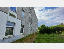 Außenansicht eines mehrstöckigen, grauen Gebäudes mit rechteckigen Fenstern und vertikal strukturierten Fassadenelementen, umgeben von einer Wiese und Büschen im Vordergrund. Der Himmel ist teils bewölkt, und im Hintergrund sind vereinzelte Bäume sichtbar.