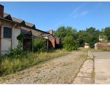 Ehemaliges Industrie- oder Lagergebäude mit weißer Fassade und Flachdach, teilweise von Pflanzen überwuchert. Kopfsteinpflaster- und Betonwege führen durch eine überwachsene, ländliche Umgebung mit Bäumen im Hintergrund.