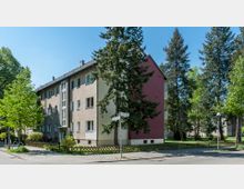 Mehrfamilienhaus mit drei Etagen und schlichtem Design, teils in beiger und teils in bordeauxroter Fassade gestrichen, umgeben von Bäumen, Grünflächen und einem niedrigen Holzzaun. Im Vordergrund eine Straßenkreuzung mit Straßenschildern und Gehwegen.