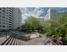 Wohngebiet mit mehreren hohen, weißen Wohnblocks im Hintergrund, umgeben von Bäumen und Grünflächen. Im Vordergrund befindet sich ein kleiner Sandspielplatz mit einer Betoneinfassung, angrenzend an eine Treppe mit bemalten Geländern.