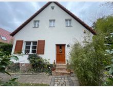 Frontansicht eines kleinen Einfamilienhauses mit weiß verputzter Fassade und steilem Satteldach mit roten Ziegeln. Zu sehen sind Holzfensterläden, eine hölzerne Eingangstür mit kurzen Backsteinstufen sowie ein Vorgarten mit Sträuchern, Bambus und Pflanzkübeln entlang eines gepflasterten Weges.