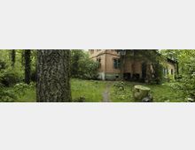 Backsteingebäude im Grünen, umgeben von dichtem Wald und wildem Gras; im Vordergrund ein Baumstamm und ein schmaler Pfad, der zum Gebäude führt. Eine alte Straßenlaterne steht neben dem Weg, Fensterläden am Gebäude sind grün gestrichen.