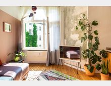 Wohnzimmer mit Holzboden und modernem Dekor. Ein großes, geöffnetes Fenster mit transparenten Vorhängen zeigt eine von Efeu bewachsene Hausfassade. Das Zimmer ist mit einem Flachbildfernseher, einer großen Zimmerpflanze, einem Sofa und einem bunten Teppich ausgestattet.