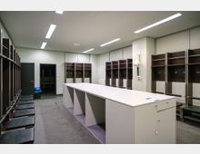 Umkleideraum mit dunklen Holzgarderoben, Haken und Sitzbänken an den Seitenwänden. In der Mitte steht ein großer, weißer Tisch, die Beleuchtung erfolgt durch Deckenleuchten.