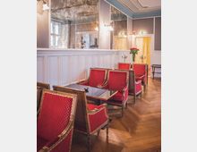 Eleganter Raum mit roten, gepolsterten Stühlen um einen kleinen Glastisch, Holzboden und einer weißen Wand mit Spiegelpanelen. An der Wand hängen klassische Wandlampen, und im Hintergrund steht ein Tisch mit Rosen und Kerzenhaltern.