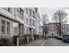 Wohnstraße mit mehrstöckigen Altbauten, teils in heller Fassade und teils mit roten Backsteinelementen. Die Straße ist gepflastert, gesäumt von geparkten Autos und Bäumen, mit kleinen Vorgärten und Metallzäunen vor den Gebäuden.