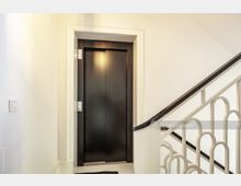 Flur eines Treppenhauses mit weiß gestrichenen Wänden; links ein schwarzer Aufzug mit moderner Tür, rechts ein Teil eines Treppengeländers mit geschwungenen Mustern.