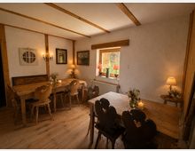 Gemütlicher Gastraum im Landhausstil mit Holzboden, sichtbaren Deckenbalken und mehreren massiven Holztischen mit geschnitzten Stühlen. An den verputzten Wänden hängen Bilder und Lampen; ein kleines Fenster mit Blick auf ein Holzgebäude lässt Tageslicht herein.