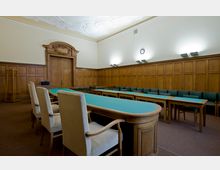 Ein historischer Sitzungssaal mit Wänden aus Holzvertäfelung, einer Decke mit dekorativen Stuckverzierungen und einer großen hölzernen Tür mit ornamentalem Rahmen. Im Raum stehen Tische mit grüner Oberfläche und gepolsterte Stühle; die Beleuchtung erfolgt durch Wandlampen.