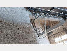 Industrieartiger Innenraum mit Sichtbetonwänden, Ziegelmauerwerk und freiliegenden Metalltreppen sowie Geländern. Im unteren Bereich sind große Fenster mit einfallendem Tageslicht zu sehen.