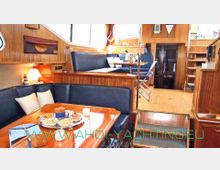 Innenraum einer Yacht mit holzgetäfelten Wänden und glänzendem Holzboden. Im Vordergrund befindet sich eine Sitzgruppe mit einem Esstisch, im Hintergrund sind erhöhte Sitzbänke, Treppen und mehrere Regale zu sehen.