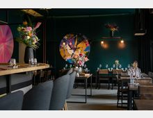Modernes Restaurant mit Holz- und Metalldesign, dunklen Wänden und einer Theke mit gepolsterten Barhockern. Der Raum ist dekoriert mit abstrakter Wandkunst, Blumenarrangements und warmen Beleuchtungselementen.