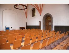 Ein heller Veranstaltungsraum mit Reihen aus hellbraunen Holzstühlen, einer weißen Wand, einem Rednerpult und einem Klavier. Die Architektur zeigt eine Gewölbedecke mit rot-orangefarbenen Bögen und eine Holztür mit einem steinernen Türrahmen.