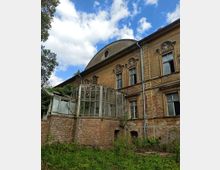 Fassade eines alten, verfallenen Gebäudes mit bröckelndem Putz und kunstvoll verzierten Fenstern. Im Vordergrund befindet sich ein einst verglaster Wintergarten mit beschädigten Glasscheiben und einer Backsteinbasis, umgeben von überwucherter Vegetation unter blauem Himmel mit Wolken.