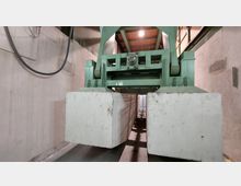 Industrieller Innenraum mit einer großen grünen Maschinenkonstruktion, die zwei massive weiße Blöcke hält. Wände aus Beton umgeben die Szene, und eine Kabelverbindung ist an der Wand sichtbar.