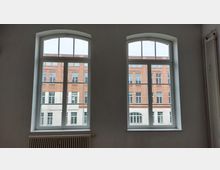 Zwei große, bogenförmige Fenster in einem weißen Innenraum bieten den Blick auf die Fassade eines gegenüberliegenden roten Backsteingebäudes mit weißen Fensterrahmen und Dekorelementen. Unter den Fenstern ist ein Heizkörper sichtbar.
