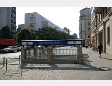 Eingang zum U-Bahnhof Strausberger Platz in Berlin, umgeben von breiten Gehwegen, Wohngebäuden im Hintergrund und einer Straße mit parkenden sowie fahrenden Fahrzeugen. Rechts im Bild sind Geschäfte, darunter eine Bäckerei, sichtbar.