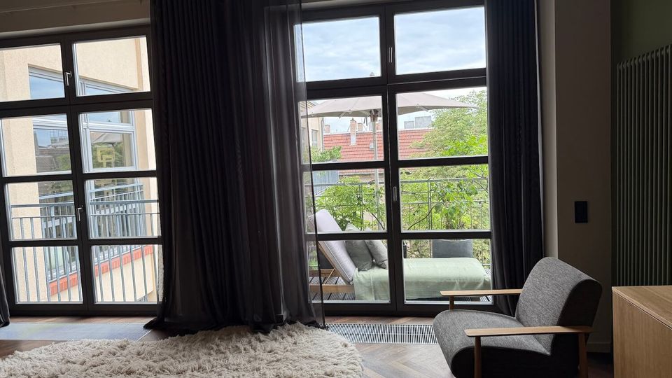 Modern bedroom with herringbone wood flooring, a round shag rug, and an upholstered chair beside floor-to-ceiling glass doors with dark curtains. The doors open to a balcony with outdoor seating and greenery, with neighboring buildings visible outside.