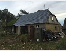 Ein altes Gebäude mit kombinierten Dachflächen, teils mit grauen Ziegeln und teils mit grünem Moos bewachsen. Der Außenbereich ist von verwilderter Vegetation umgeben, während rechts neben dem Haus Gegenstände und Baustellenmaterialien unsortiert gestapelt sind.