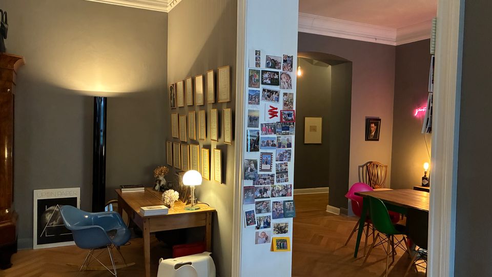Blick in eine Wohnung mit hohen Decken, Stuckleisten und Parkettboden: links ein kleiner Arbeitsplatz mit Holztisch, Stuhl und Wandrahmen, in der Mitte ein Türrahmen mit Fotocollage. Rechts schließt ein Essbereich mit Holztisch, bunten Stühlen und gedämpfter Beleuchtung an, dahinter ein Flur mit Rundbogen.