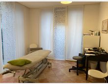 Behandlungsraum mit einem höhenverstellbaren Therapieliege in der Mitte, links ein grünes Kissen darauf. Im Hintergrund weiße Vorhänge mit dekorativem Muster vor Fenstern, rechts ein Schreibtisch mit Büroartikeln und ein Schreibtischstuhl. Fliesenboden in warmen Tönen.