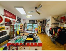 Innenraum eines hobbyartigen Werkstatt- und Sammlerraums mit Holzfußboden, Deckenventilator und großen Fenstern; an den Wänden hängen Vintage-Auto- und Tankstellen-Schilder, Teile und Dekoration. Im Vordergrund steht ein Tischkicker, daneben Vitrinen und Regale mit Gegenständen sowie ein Kleiderständer und diverse Fahrzeugteile.