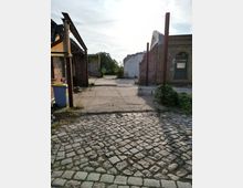 Ansicht eines teilweise verfallenen Gebäudekomplexes mit gemauerten Wänden, beschädigten Dächern und rostigen Stahlstreben. Im Vordergrund ist ein Kopfsteinpflaster zu sehen, im Hintergrund dichter Bewuchs und ein heller Himmel.