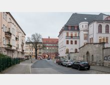Straßenszene in einer Wohngegend mit mehrstöckigen Gebäuden im Altbaustil, darunter ein großes, weißes Gebäude mit roten Fenstern und einem grauen Schieferdach. Am Straßenrand sind Autos geparkt, und ein Baum ohne Laub steht auf dem Gehweg; eine Mauer mit Graffiti ist rechts sichtbar.