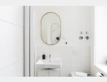 Modernes, helles Badezimmer mit weißen Fliesenwänden, einem ovalen Spiegel mit goldenem Rahmen, einem weißen Waschbecken sowie einer schwarzen Seifenspenderflasche. Im Hintergrund ist eine Duschkabine zu erkennen.