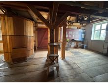 Innenraum einer historischen Mühle mit Holzboden, Holzbalken und einer großen hölzernen Trommel. Im Hintergrund sind eine rote Holztür sowie Informationstafeln und Ausstellungsstücke erkennbar. Ein Fenster lässt Tageslicht in den Raum.