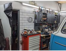 Werkstatt mit weiß gefliesten Wänden, roten und grauen Werkzeugschränken, sowie einer Arbeitsfläche mit Werkzeugen und Utensilien. Rechts steht teilweise ein blau-weißer Kleinbus, und links ist eine offene Tür zu einem weiteren Raum sichtbar.
