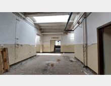 Ein verlassener, industrieller Innenraum mit abblätternder Farbe an den Wänden, sichtbaren Rohren an der Decke und einem Schild mit der Aufschrift "E-Werkstatt" über einer Tür. Der Boden ist mit Schutt und Staub bedeckt, und Tageslicht fällt durch Deckenfenster ein.