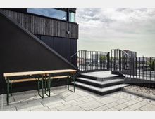Eine moderne Dachterrasse mit hellgrauem Pflasterboden, einer schwarzen Treppe mit Metallgeländer und einer Holz-Sitzbank. Im Hintergrund ist ein Ausblick auf eine urbane Skyline und einzelne Grünflächen zu sehen.