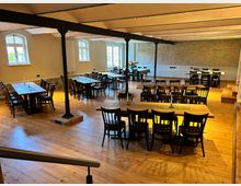 Innenansicht eines Veranstaltungsraums mit Holzboden, mehreren rechteckigen Holztischen und schwarzen Stühlen. Der Raum hat helle Wände, große Fenster mit Rundbögen, freiliegende Stahlstützen und eine Backsteinwand im Hintergrund.