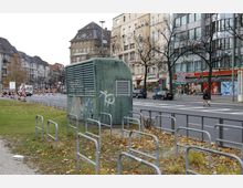 Straßenszene in einer Stadt mit mehrstöckigen Wohn- und Geschäftshäusern im Hintergrund, einem kleinen grünen Gebäude mit Lüftungsgittern und Graffiti im Vordergrund sowie abgestellten Fahrradständern. Herbstliche Bäume und trockenes Laub prägen die Umgebung.