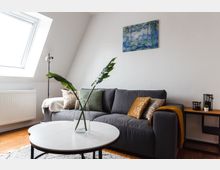 Helles Wohnzimmer mit einer grauen Couch, die mit bunten Kissen dekoriert ist. Ein runder, weißer Couchtisch mit einer Vase und großen grünen Blättern steht davor; im Hintergrund ein schräges Dachfenster, ein abstraktes Gemälde an der Wand und ein kleiner Holz-Beistelltisch mit Dekoobjekten.