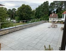 Große Terrasse mit grauen Steinfliesen, umgeben von einer niedrigen Brüstung aus Naturstein mit Metallgeländer. Im Hintergrund befinden sich grüne Bäume, eine Rasenfläche und einzelne Häuser mit weißen Fassaden und roten Dächern.
