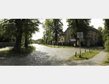 Pflastersteinstraße in einer ländlichen Wohngegend mit alten Häusern im Hintergrund, gesäumt von Bäumen. Ein Verkehrsschild weist auf eine 30-km/h-Zone hin, und ein Fußweg verläuft entlang der Straße.