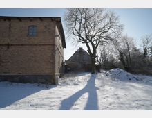 Verschneiter Hofbereich mit einem großen, kahlen Baum in der Mitte, dessen Schatten über den Schnee fällt. Links steht ein zweigeschossiges Backsteingebäude, im Hintergrund ein weiteres Wirtschaftsgebäude mit Satteldach, umgeben von winterlichen Bäumen und Sträuchern.