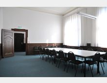 Seminarraum mit großem rechteckigem Tisch und schwarzen Stühlen in einer hell beleuchteten Umgebung. Holzvertäfelungen an den Wänden, ein offener Holztürrahmen sowie bodenlange weiße Vorhänge vor großen Fenstern sind sichtbar.