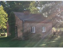 Kleines Backsteinhaus mit Satteldach in einem Garten, umgeben von Bäumen. Efeu wächst an der linken Hauswand, und im Vordergrund ist eine Grünfläche mit einem Brunnen und einem hölzernen Gartentisch sichtbar.