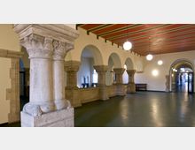 Halle mit historischen Steinsäulen, verzierten Kapitellen und Rundbögen, Decke in warmen Rottönen mit Streifenmustern, beleuchtet von kugelförmigen Lampen, seitlich eingelassene Fenster und Sitzbänke.