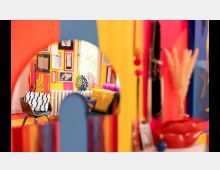 Ein bunt eingerichteter Raum mit leuchtend roten, gelben und blauen Wänden, dekoriert mit Kunstwerken und gerahmten Bildern. Im Vordergrund steht ein Stuhl mit gemustertem Kissen, während farbige Kissen und Trockenblumen die dekorative Atmosphäre ergänzen.