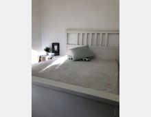 Ein schlicht eingerichtetes Schlafzimmer mit einem weißen Holzbett, dessen Kopfteil aus vertikalen Streben besteht. Auf dem Bett liegt eine helle Decke sowie zwei Dekokissen, und daneben befindet sich ein kleiner geflochtener Nachttisch mit einer Pflanze und einem kleinen Bilderrahmen.